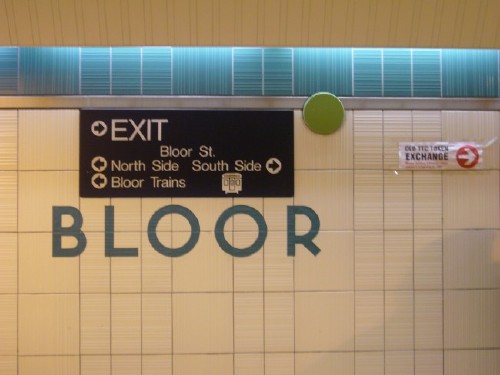 TTC font in tile wall reads BLOOR. Sign in Helvetia with arrows sits over it. A green dot is attached to the wall. Taped-up paper sign eads OLD TTC TOKEN EXCHANGE