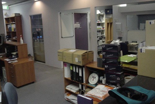 Wooden L-shaped desk in grey-carpeted office holds a few boxes and binders. Green hat sits in foreground