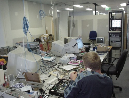 Blond man chats on telephone in a room with bare drywall, cables hanging from the ceiling, and computers and electronics strewn on a long table and piled on the floor