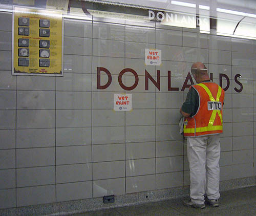 Donlands station has WET PAINT signs as man in safety vest works on the tile wall
