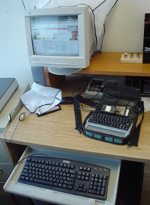 Monitor, keyboard, headphones,  Braille notetaking device, and tape player sit on desk