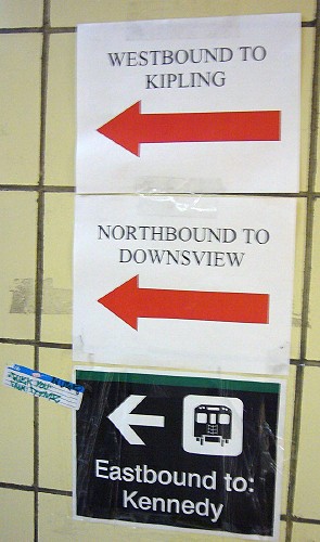 Two ink-jet-printed signs with red arrow and black Times type on white; one sign with a green line across the top and white ‘Helvetica’ type and arrow
