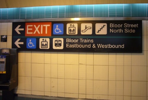 Sign on wall in ‘Helvetica’ has nine words, two arrows, one EXIT logotype, and seven pictographs, two of which are repeated