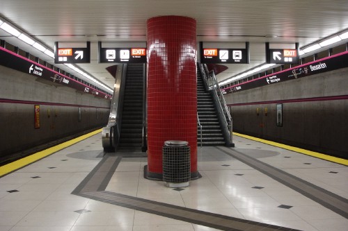 Subway platform has wayfinding tiles leading around bright-red-tiled pillar. ‘Helvetica’ signs hang from roof over escalators