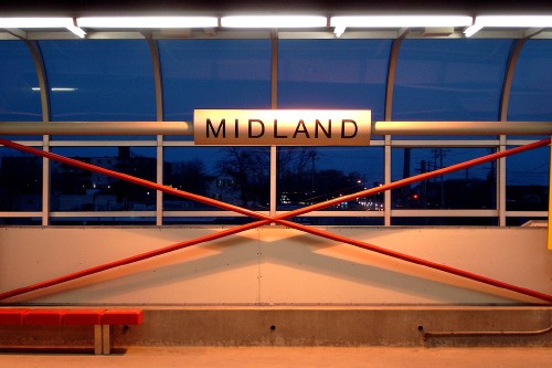 Against a dusky sky, a curved metal board over criscrossing members reads MIDLAND in Helvetica