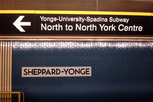 Sign on blue tile wall reads SHEPPARD-YONGE on a metal plate. Overhead sign in ‘Helvetica’ reads ← Yonge-University-Spadina Subway North to North York Centre