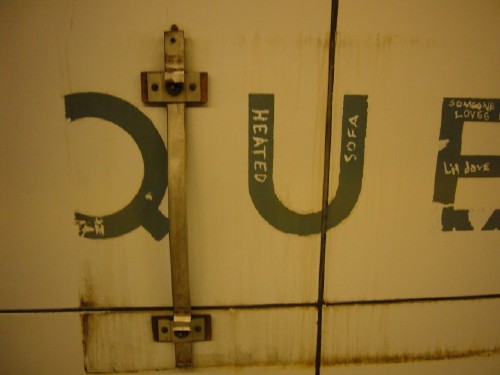 Metal strap is attached to wall amid broken-up letters Q U E in which graffiti is written