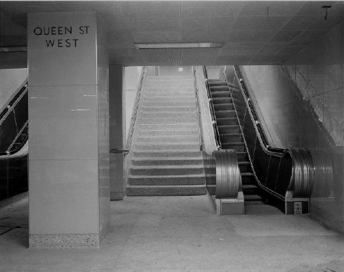 Spare subway-station concourse by escalators uses large glossy tiles and reads QUEEN ST WEST