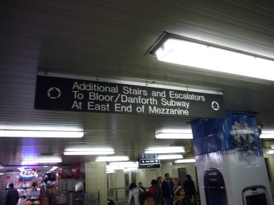Ceiling-mounted sign reads Additional Stairs and Escalators to Bloor/Danforth Subway At East End of Mezzanine