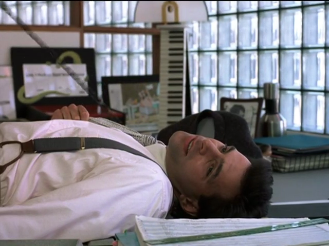 Michael, in shirt and suspenders, lying down on office desk with a wan smile