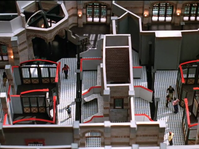 Overhead view of diorama shows mezzotint-patterned flooring, office cubicles with red-topped walls, this time with models of people