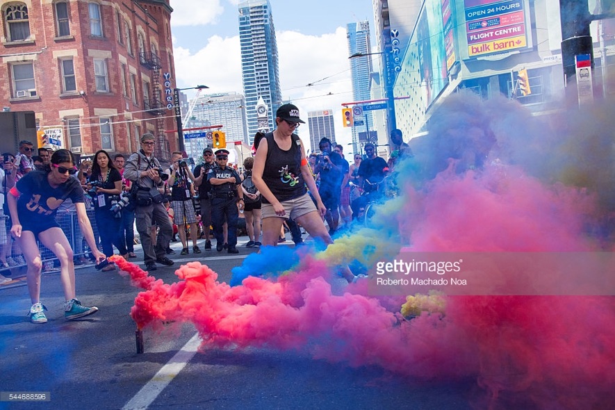 Humanoid female sets down a canister, already emitting pink smoke, while another humanoid female and many other people, including a cop and a photographer, watch