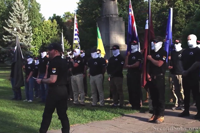 Man without face covering walking before rank of men in identical branded polo shirts and white face coverings
